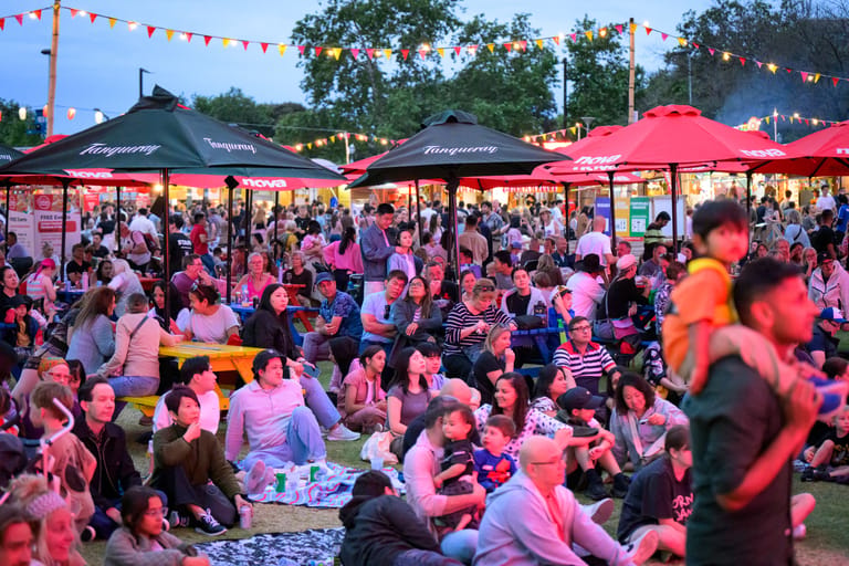 Crowds gather under pink-lit umbrellas and marquees at an outdoor evening festival or night market event.
