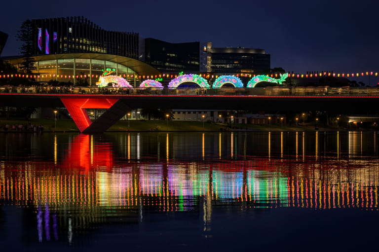 Vibrant coloured lights illuminate Adelaide's riverbank at night, reflecting across the water below historic buildings.