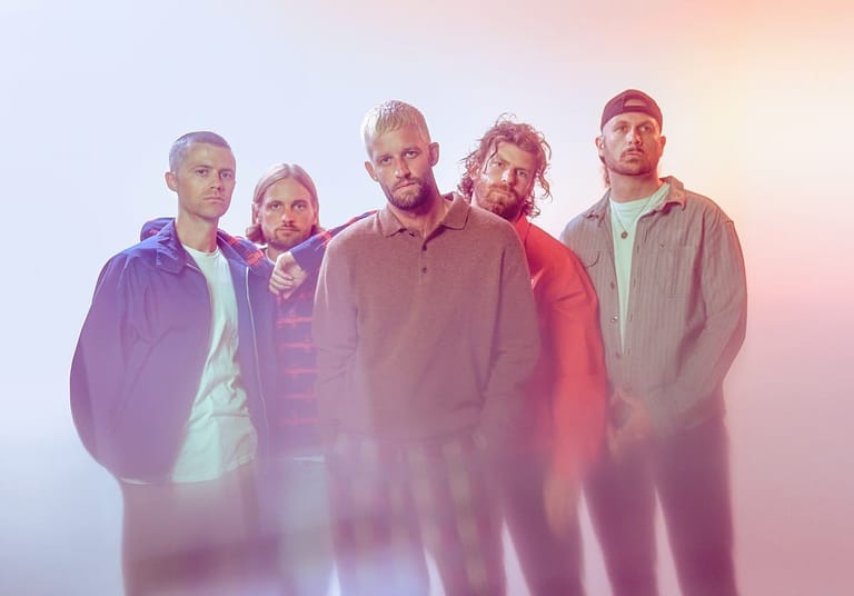 Five band members stand together against a soft pink and purple gradient background lit by atmospheric stage lighting.