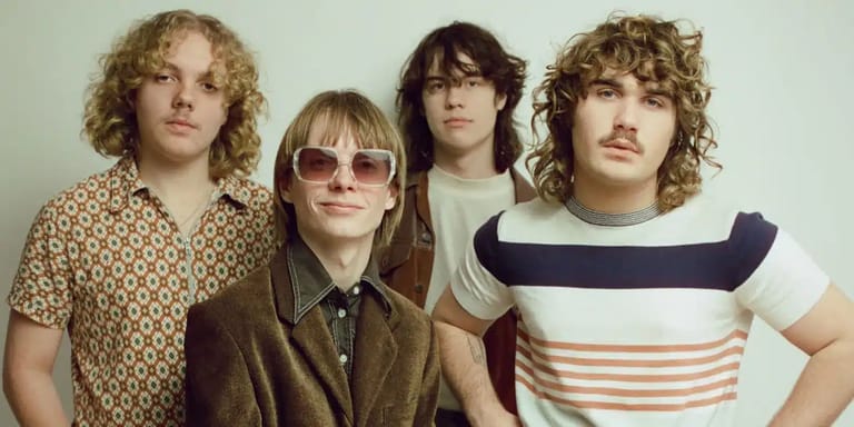 Four band members with curly and straight hair pose together against a neutral background.