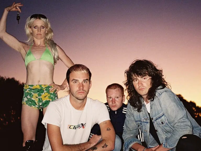 Four band members pose against a twilight sky, one shirtless in colourful shorts elevated above the others.
