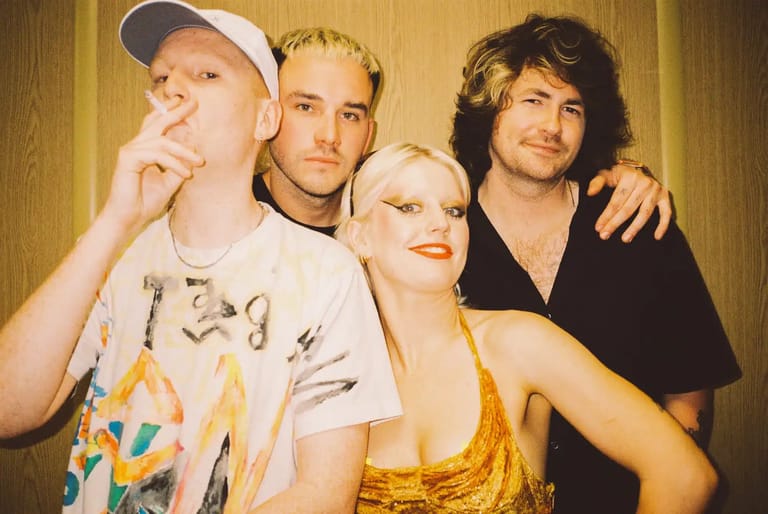 Four musicians pose together against a golden textured backdrop, with one wearing a white cap and graphic tee, another in a black shirt, and a woman in a gold sequined dress.