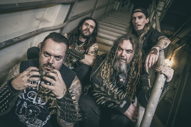 Five tattooed band members pose together in a dimly lit industrial setting with metal stairs.