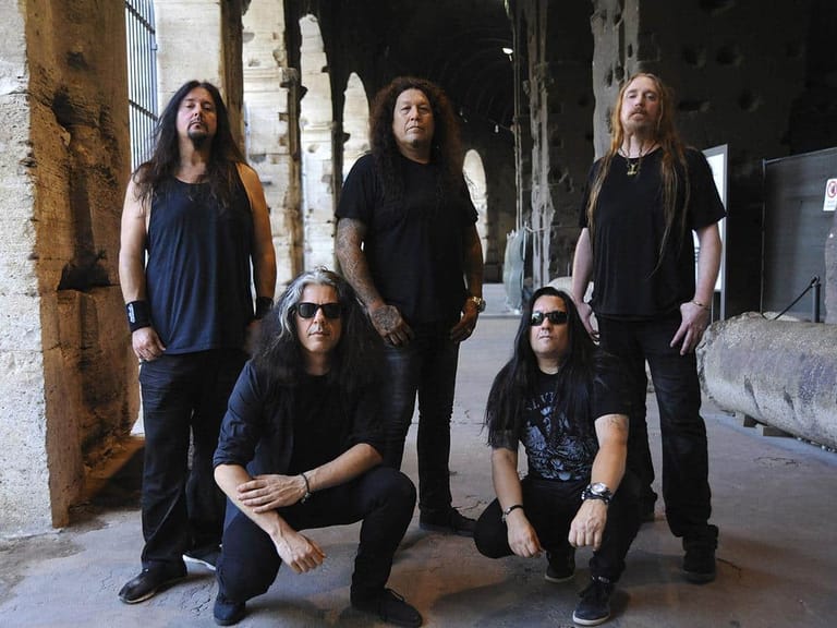 Six musicians in dark clothing pose together in an industrial warehouse setting with concrete walls.
