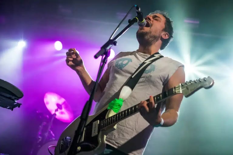 Male musician singing into microphone whilst playing electric guitar under purple and green stage lighting.