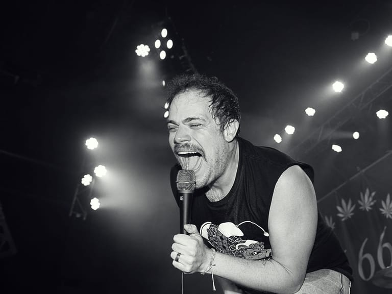 Male performer singing into microphone on stage, lit by bright spotlights in black and white.
