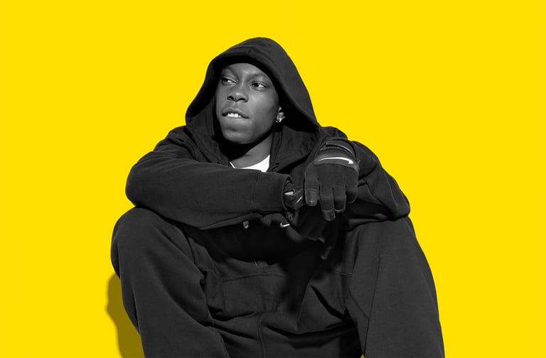 Person in black hoodie and jacket seated against bright yellow background, looking upward.