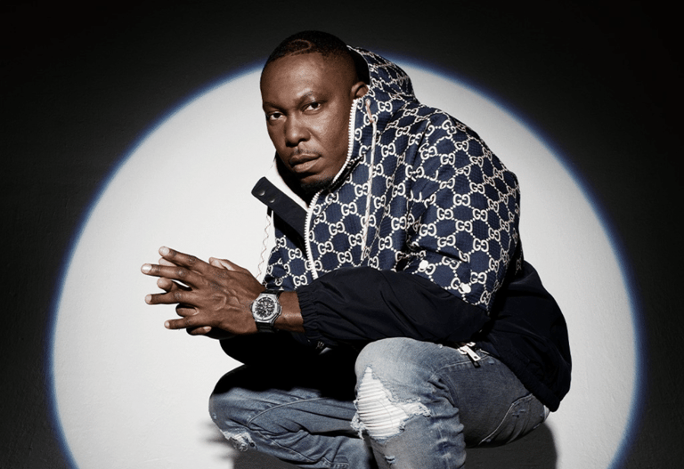Man in designer monogrammed jacket crouches against white circular backdrop, wearing watch and rings.