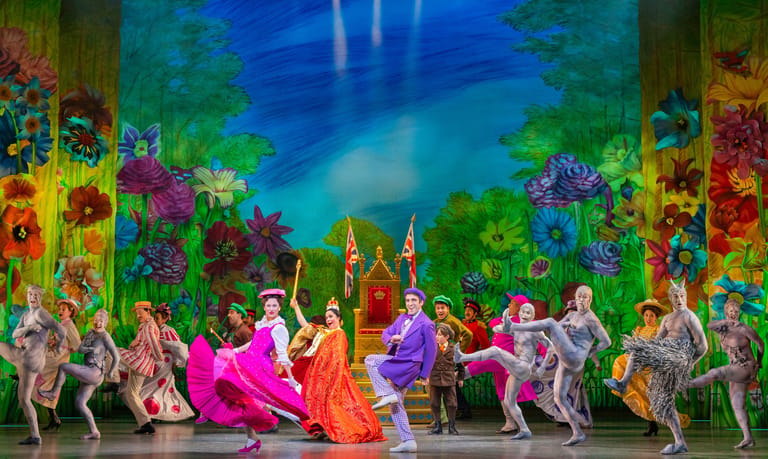 Dancers in vibrant costumes perform on stage before a colourful painted backdrop of flowers and a blue sky.