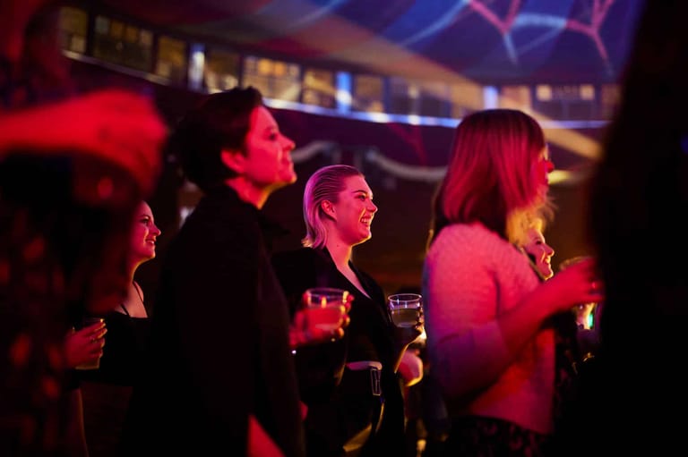Group of people holding drinks in a dimly lit venue bathed in red stage lighting.