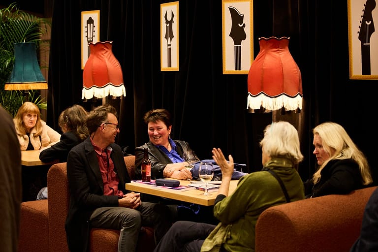 Panel of five people seated around a table during a theatre discussion or Q&A session, with orange pendant lights overhead.