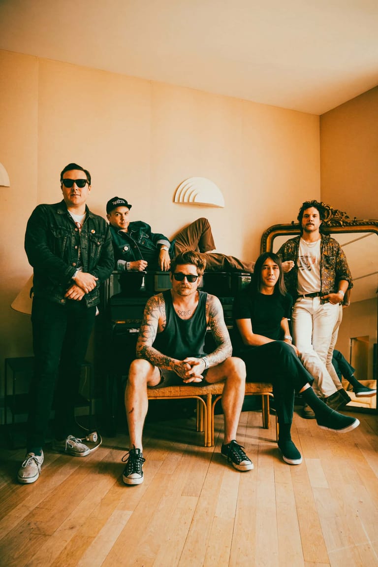 Five band members posed in a warm-lit living room, wearing casual dark and neutral clothing.