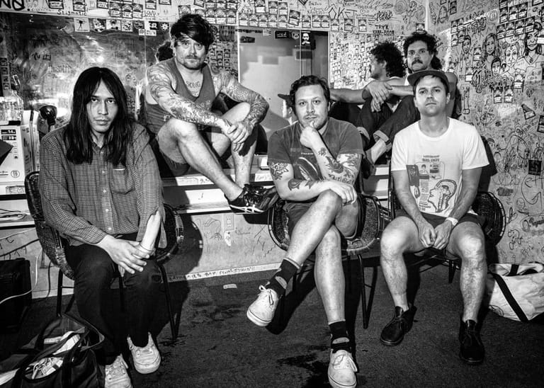 Six people sit together in a cluttered black-and-white interior space decorated with posters and memorabilia.