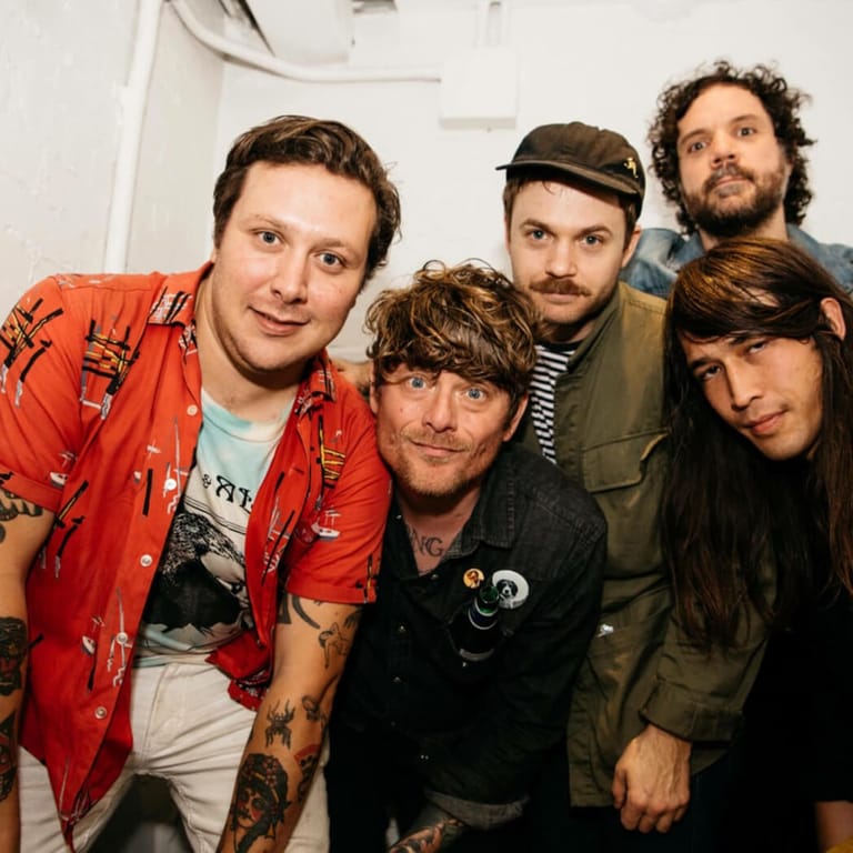 Five band members posing together indoors, wearing casual and colourful clothing including a red shirt and various hats.