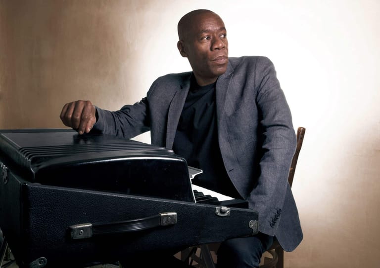 Man in grey blazer seated at grand piano against neutral backdrop.