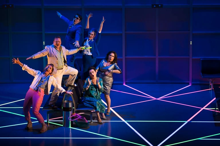 Performers in colourful costumes strike dynamic poses on an illuminated geometric stage set with blue lighting.