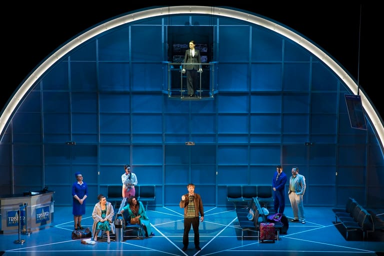 Cast members positioned across a blue-gridded stage set beneath an arched cyclorama during a theatrical production.