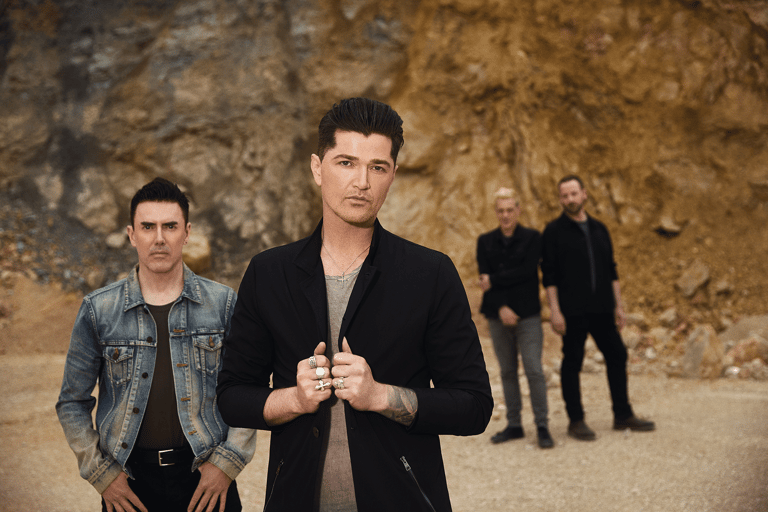 Four band members stand against a textured brown stone wall, with two in sharp focus in the foreground.