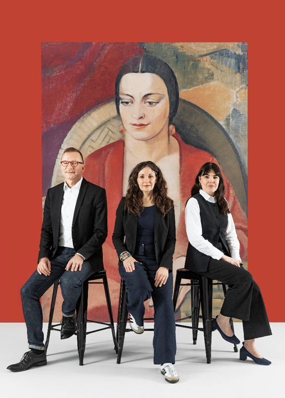 Three people seated on stools against a red wall backdrop featuring a large stylised portrait painting.