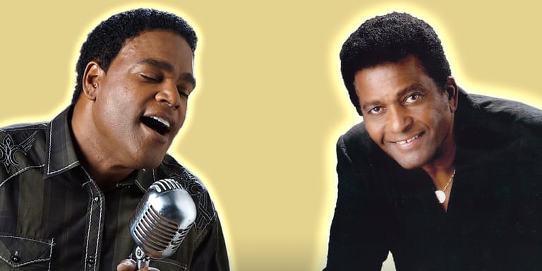 Two men in black leather and dark clothing share a microphone against a yellow background, smiling.