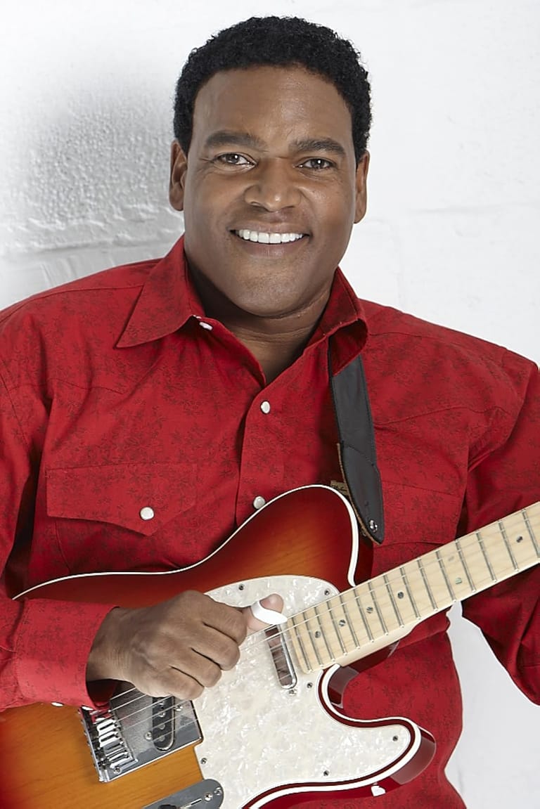 Man in red shirt smiling while holding a sunburst electric guitar against a white background.