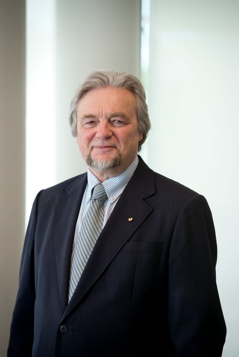 Middle-aged man with grey hair and beard wearing a dark suit and striped tie against a white wall.
