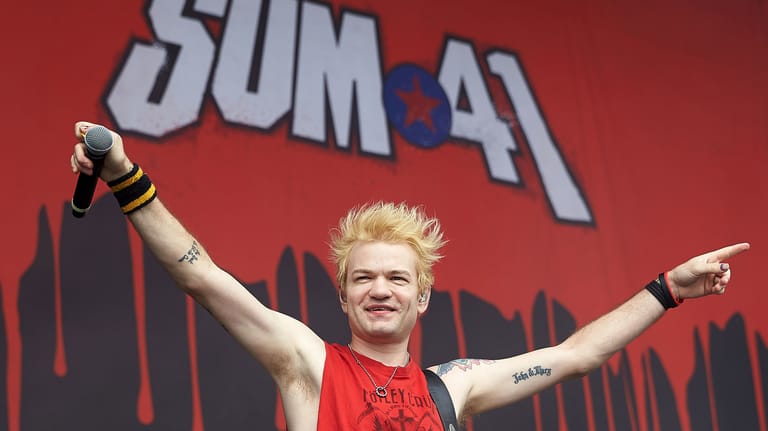 Blonde musician with raised arms performing on stage with red SUM 41 backdrop behind them.