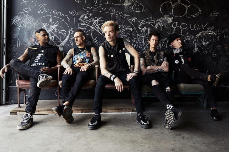 Five band members in dark clothing seated against a blackboard wall covered in sketches and writings.