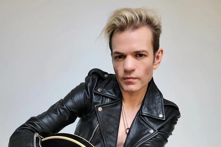 Person in black leather jacket holding an acoustic guitar against a neutral background.