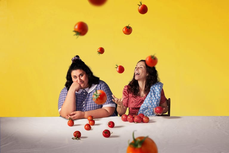 Two people sit at a white table against a bright yellow background, looking thoughtful as red apples fall around them.