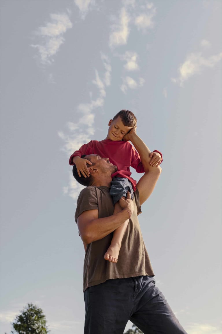 Adult holding a child aloft against a bright blue sky with scattered white clouds.