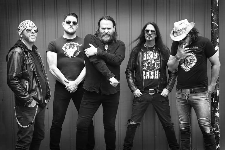 Five band members in dark clothing pose against a corrugated metal backdrop in black and white.