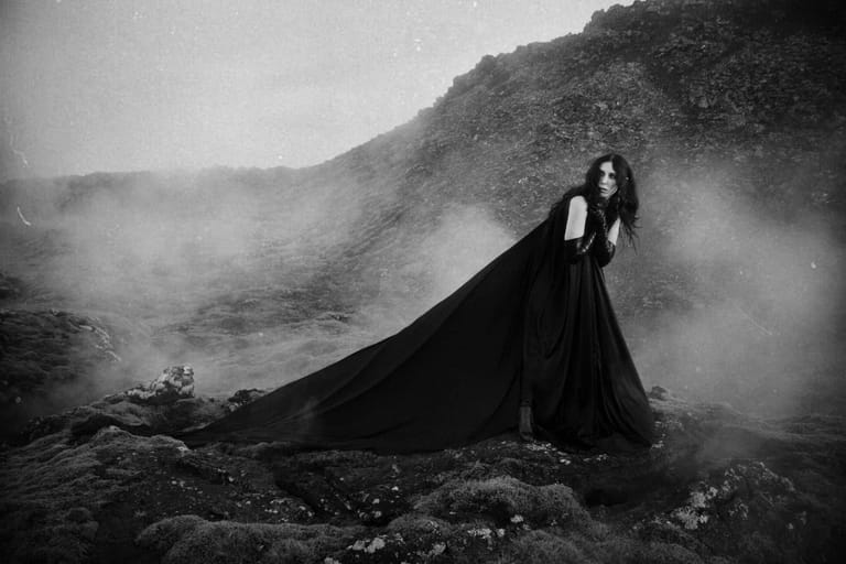 Woman in flowing black cloak standing on rocky outcrop in mist-shrouded mountainous landscape.