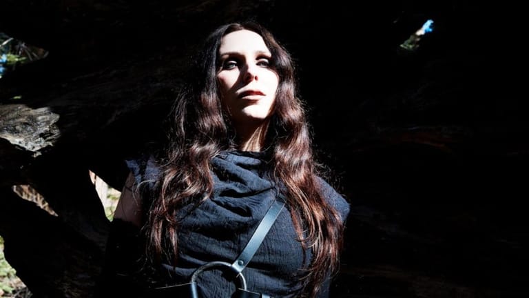 Woman with long dark hair in dark clothing against a black background with atmospheric lighting.