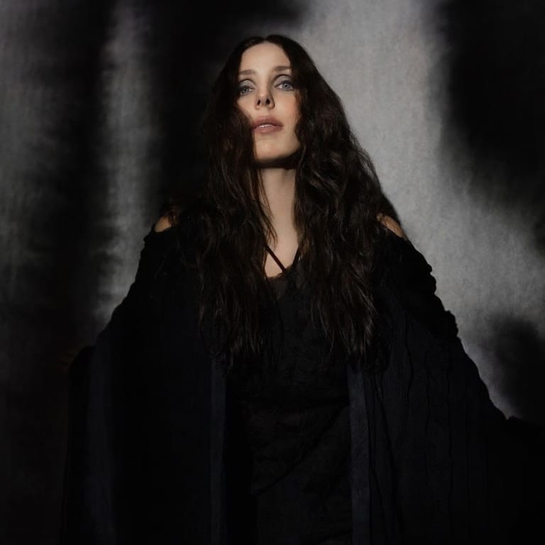 Woman with long dark hair wearing a black draped garment against a moody grey textured backdrop.