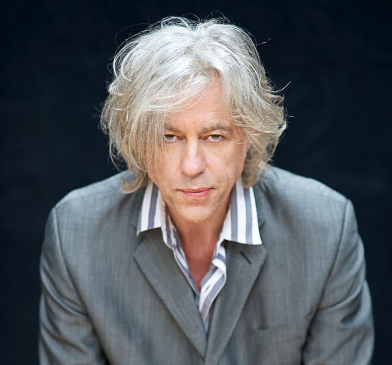 Person with white hair wearing a grey blazer and striped shirt against a dark background.