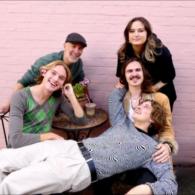 Five band members posed together against a pink brick wall, smiling at the camera.