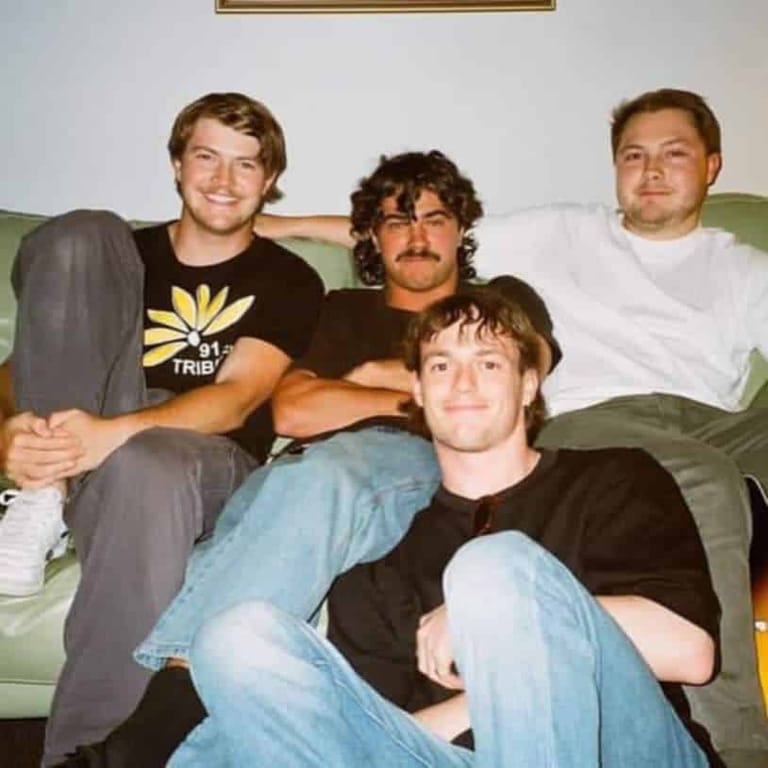 Four men posed together on a green couch in casual clothing and relaxed poses.