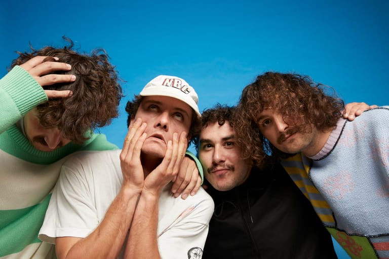Four young men with curly hair pose together against a bright blue sky, making animated expressions toward the camera.