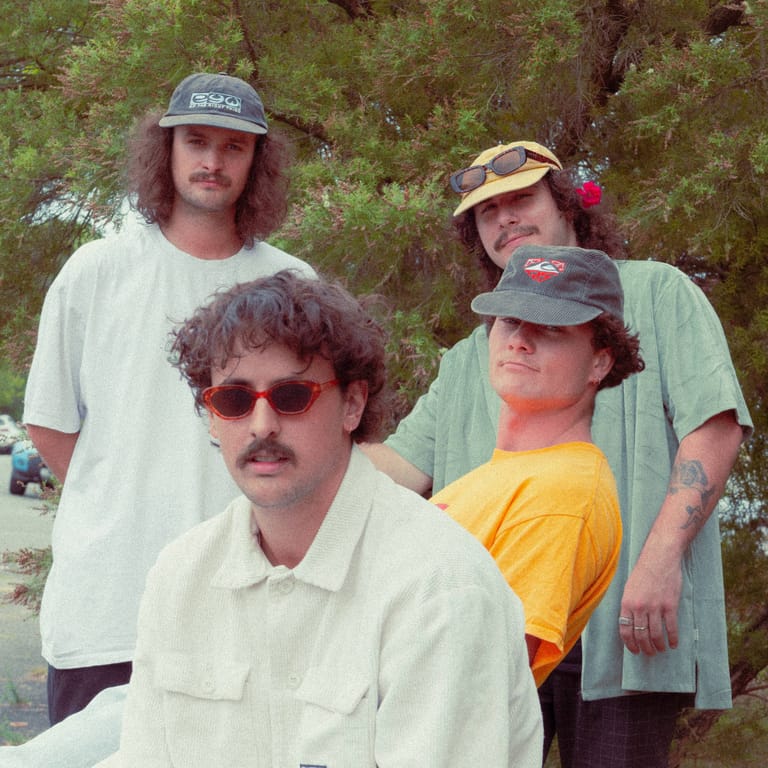 Four musicians in casual vintage-style clothing and caps pose outdoors against greenery.