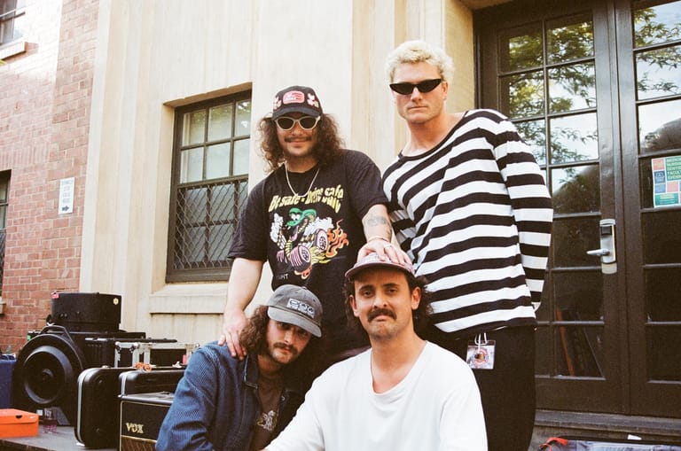 Four musicians posing together outside a brick building entrance, wearing casual clothing and sunglasses.