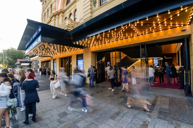 Crowds gather outside an illuminated theatre entrance on a city street at dusk.