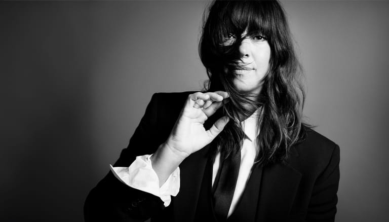 Person in black blazer and tie adjusting collar against neutral background, black and white portrait.