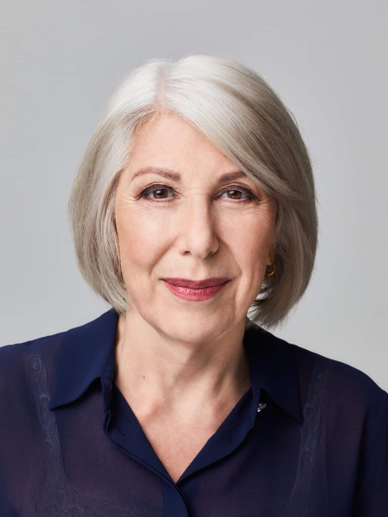 Woman with silver hair wearing a dark navy shirt against a neutral grey background.