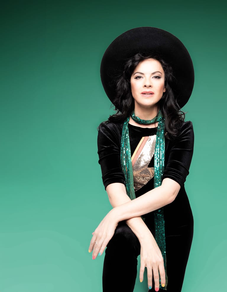Woman in black velvet jacket and wide-brimmed hat against turquoise background, arms crossed in contemplative pose.