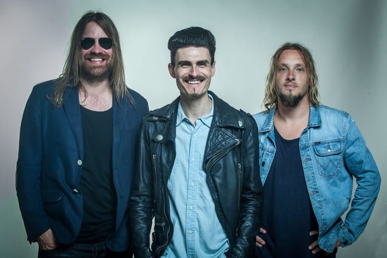 Three men with long hair and sunglasses stand against a neutral background in casual rock-style clothing.