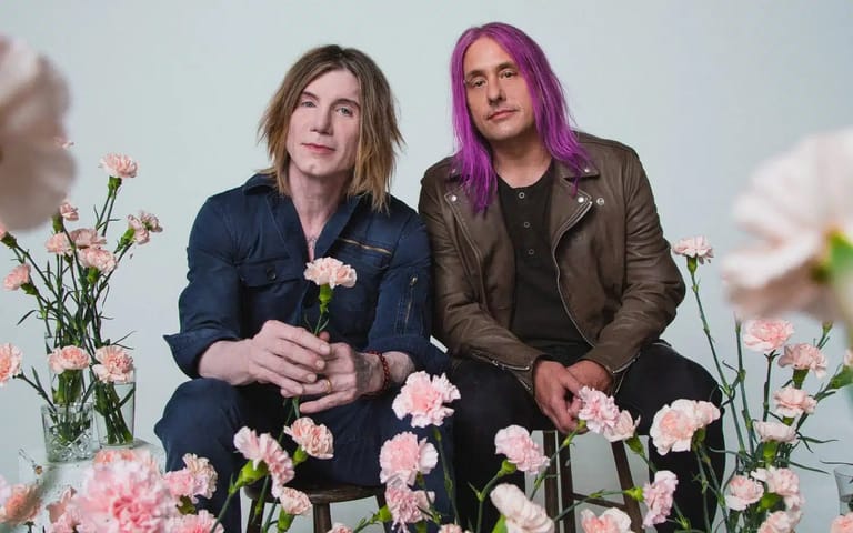 Two musicians sit among pink carnations against a white backdrop, one in a navy shirt and the other with purple-tinted hair wearing a brown jacket.