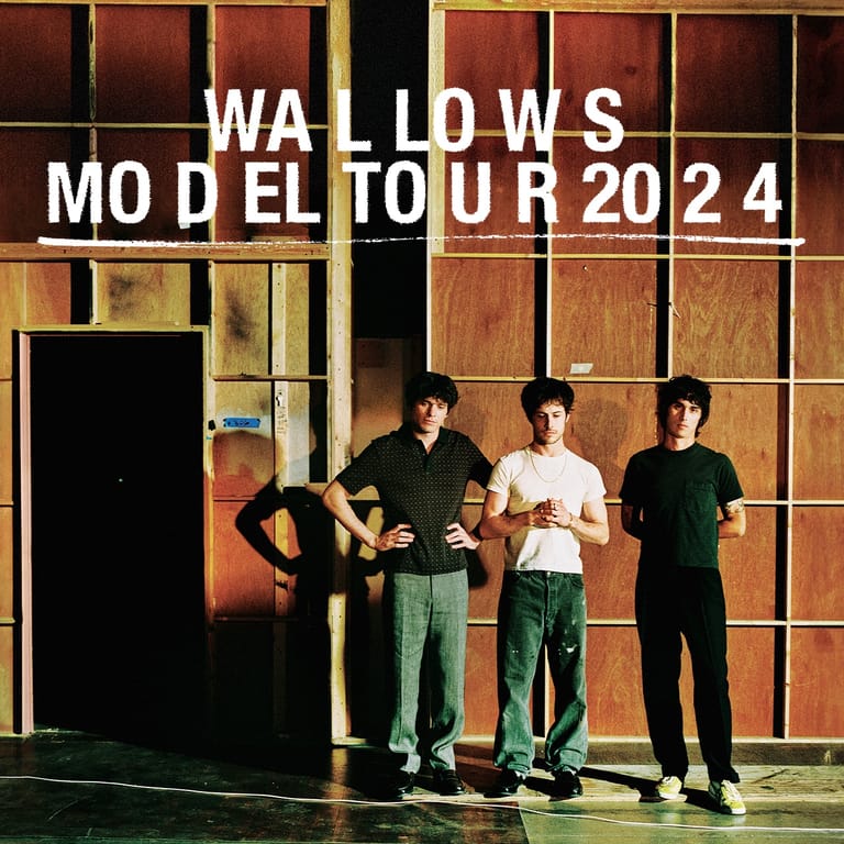 Three band members stand against a warm-toned wooden wall backdrop with "WALLOWS MODEL TOUR 2024" text overlaid.