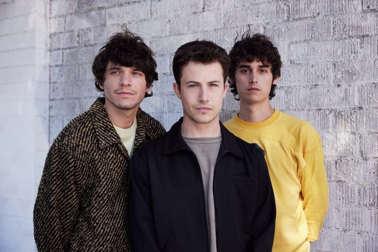 Three young men in casual layered clothing stand against a textured white brick wall.