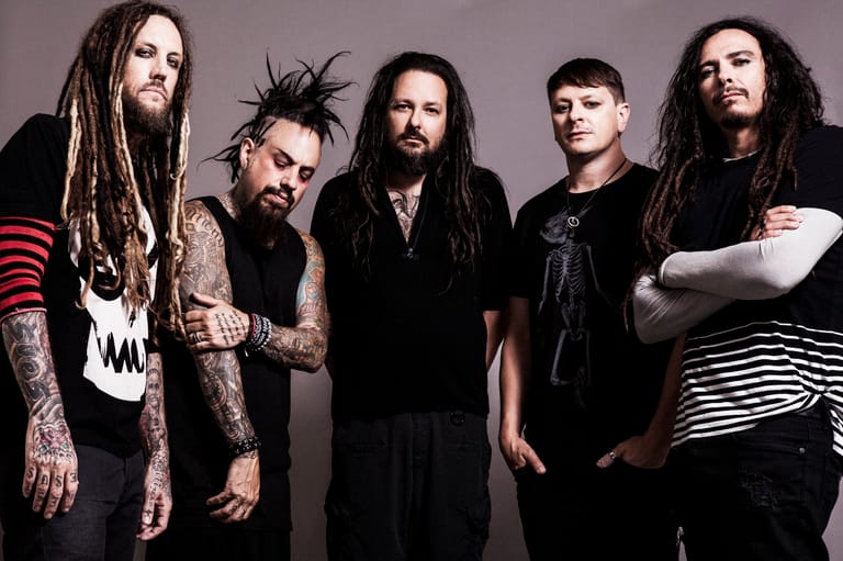 Five musicians with long dark hair and tattoos pose against a grey backdrop in band portrait format.